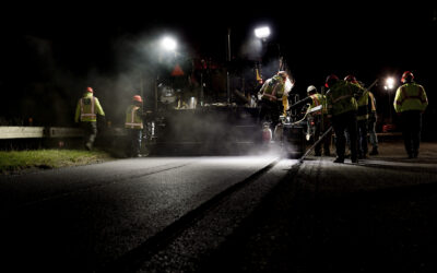 Construction crew in high-visibility jackets and hard hats work at night around heavy machinery with bright floodlights and smoke.