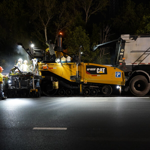 Nighttime roadwork scene with workers in safety vests and hard hats operating a yellow CAT asphalt paver beside a large dump truck.