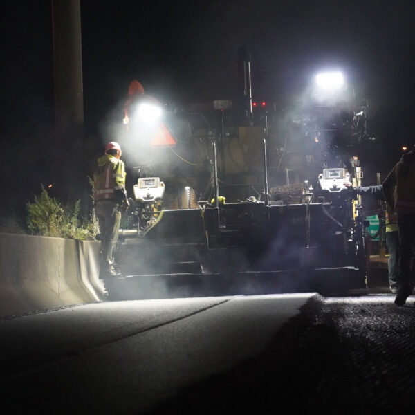 Nighttime road construction scene with workers in reflective vests and hard hats operating large machinery under bright floodlights, with a concrete barrier in the foreground and smoky air.