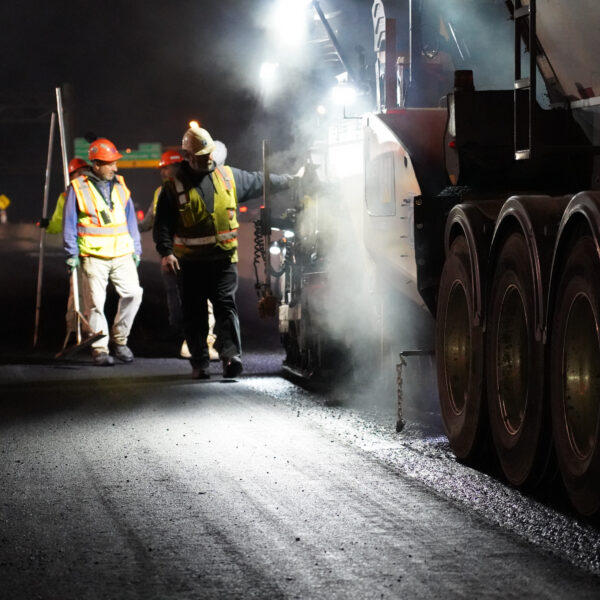 Nighttime road construction crew in high‑visibility vests and hard hats standing beside a large dump truck, with lights and steam in a dimly lit highway work zone