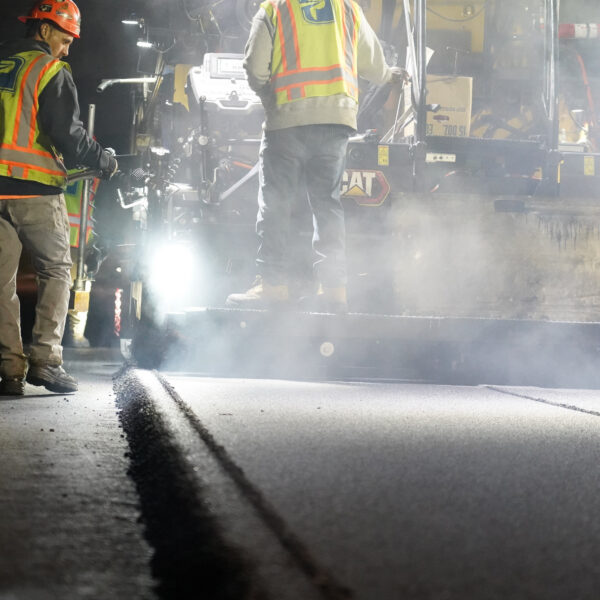 Two construction workers in neon safety vests and hard hats stand beside a large Caterpillar machine at night, surrounded by mist and dim lights.