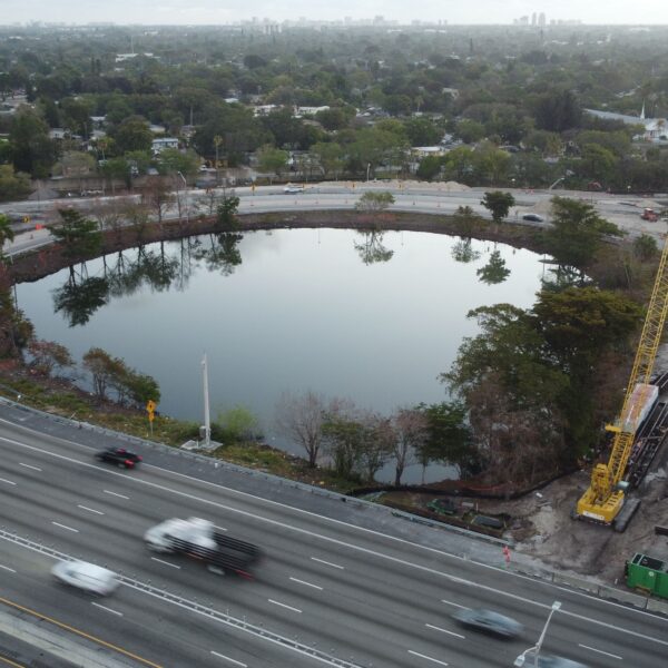 Aerial view of a highway interchange with a circular pond, trees, and active construction including a tall yellow crane and traffic cones nearby and vehicles on surrounding roads.