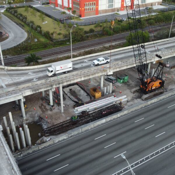 Aerial view of a highway interchange under construction, with a large crane, concrete supports, and a yellow school bus nearby beside railroad tracks in the background.