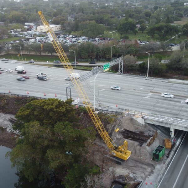 Aerial view of a multi-lane highway with a tall yellow construction crane over the roadway and a river on the left, surrounded by trees and construction equipment.
