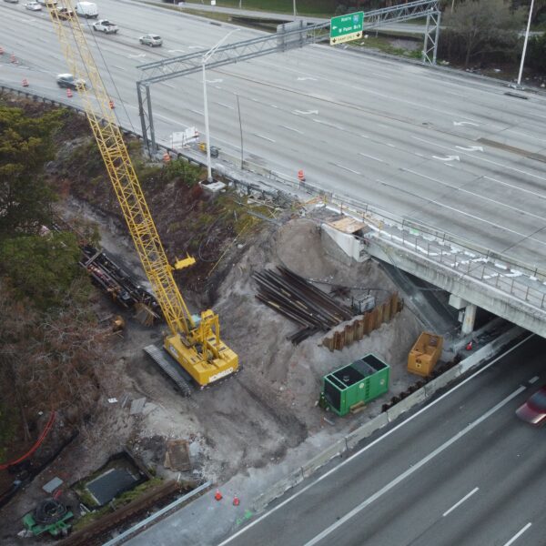 Aerial view of a construction site beside a multi-lane highway with a yellow crane, debris, and temporary barriers under an overpass, near trees and water edge.