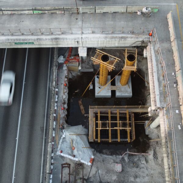 Aerial view of a construction site beneath a raised highway, showing two yellow cylindrical formwork columns with wooden supports and surrounding debris next to a busy roadway with moving cars on the left and a red car on the right.