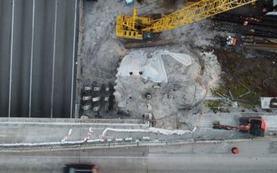 Aerial view of a construction site with a large yellow crane, dirt, and scattered materials near a roadway at the bottom of the image.