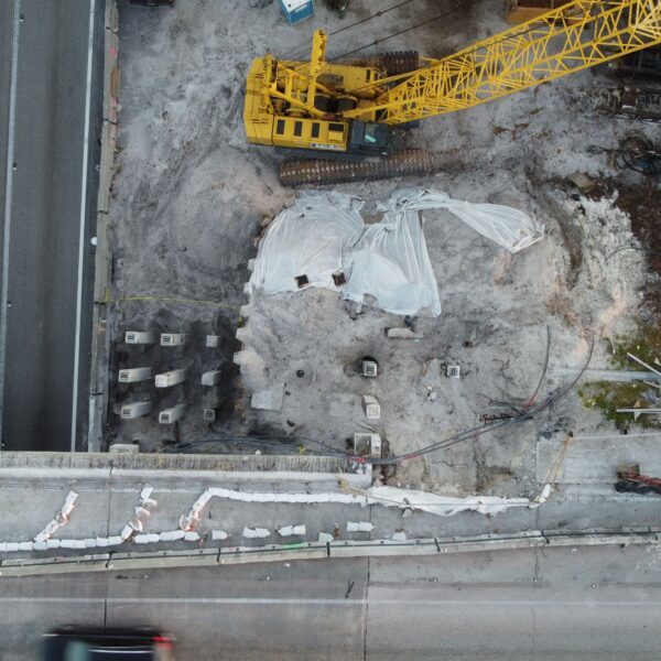 Aerial view of a construction site with a large yellow crane, dirt, and scattered materials near a roadway at the bottom of the image.