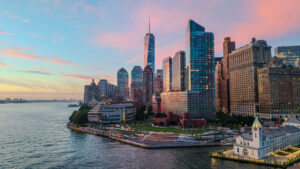 default - Posillico Inc. Manhattan skyline at sunset along the waterfront, featuring One World Trade Center and glass-skin buildings under a pink-blue sky.