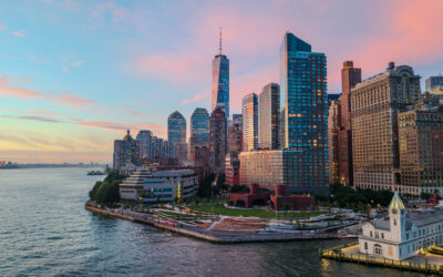Sunset skyline of downtown Manhattan with tall skyscrapers along the waterfront and a calm harbor in the foreground.