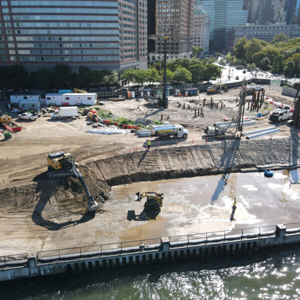 default - Posillico Inc. Urban construction site with heavy machinery and cranes along a riverfront, surrounded by tall buildings and trees.